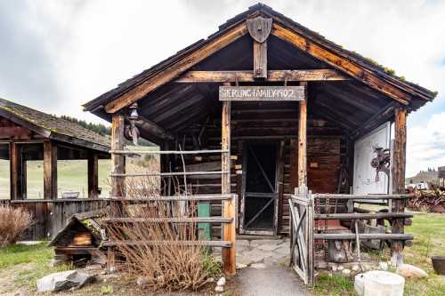 Historic wooden cabin with a sign reading "Sherling Family 1902," surrounded by rustic fencing and overgrown shrubs.