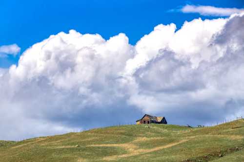 A solitary wooden house on a grassy hill under a blue sky with fluffy white clouds.