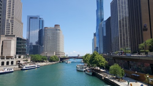 A view of the Chicago River with boats, surrounded by tall buildings and clear blue skies.