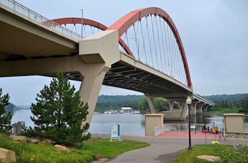A modern bridge with a red arch spans over a calm river, surrounded by greenery and a pathway.