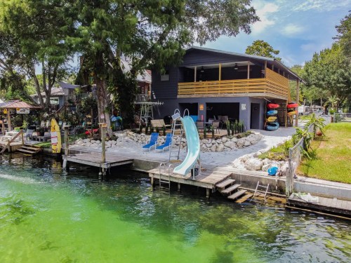 A waterfront house with a slide, surrounded by greenery and kayaks, overlooking a greenish waterway.
