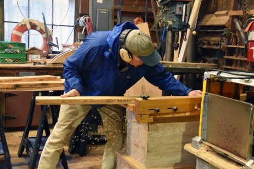 A person in a blue jacket and headphones works on wood in a workshop, using a hand tool with wood shavings around.
