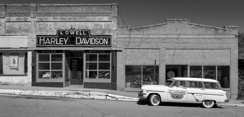 Black and white image of a vintage car parked outside a Harley-Davidson dealership in Lowell, showcasing historic architecture.