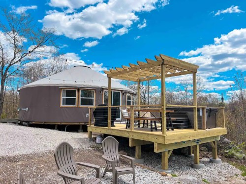 A cozy yurt with a wooden deck and outdoor seating, surrounded by trees under a bright blue sky with fluffy clouds.