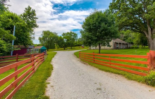 A gravel driveway lined with orange fences leads to a house surrounded by trees and greenery under a cloudy sky.