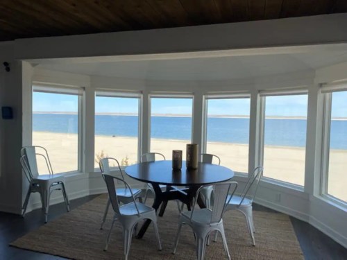 A bright dining area with a round table and chairs, overlooking a beach and ocean through large windows.