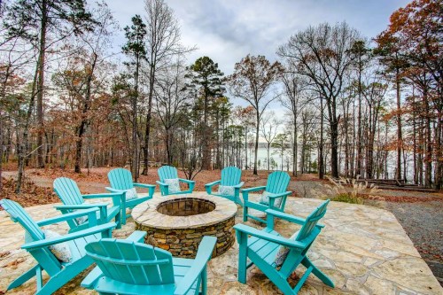 A stone fire pit surrounded by blue Adirondack chairs, set in a wooded area near a lake.