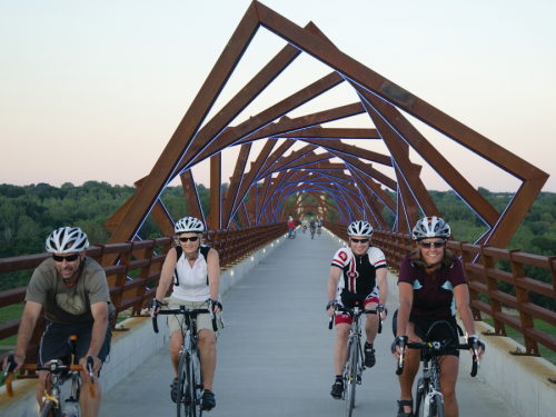 Four cyclists ride across a modern, artistic bridge with a geometric design, surrounded by greenery at sunset.