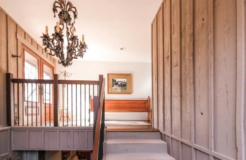 A well-lit staircase with a chandelier, leading to a landing with wooden railings and a framed painting on the wall.