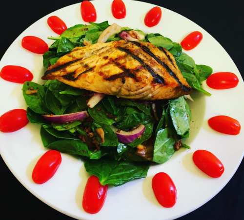 Grilled salmon on a bed of spinach, topped with red onions and surrounded by cherry tomatoes on a white plate.