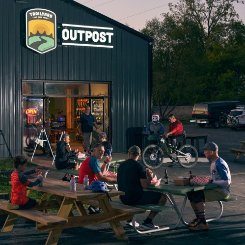 A group of people enjoying food outside a bike-themed restaurant, with a sign reading "Trail Yard Outpost" in the background.