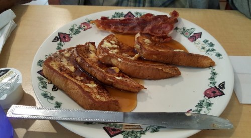 A plate of French toast drizzled with syrup, accompanied by crispy bacon strips and a knife on the side.