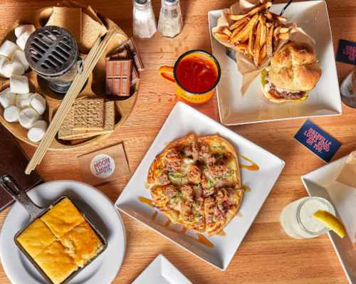 A table spread with various dishes: s'mores ingredients, sweet potato fries, a burger, and a pizza.