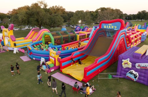A colorful inflatable playground with slides and bounce houses in a park, filled with children and families enjoying the attractions.