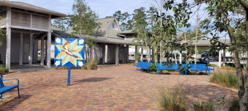 A colorful mural stands in a brick-paved courtyard surrounded by benches and trees, with buildings in the background.