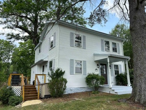 A light blue two-story house with white shutters, a front porch, and a wooden staircase surrounded by greenery.