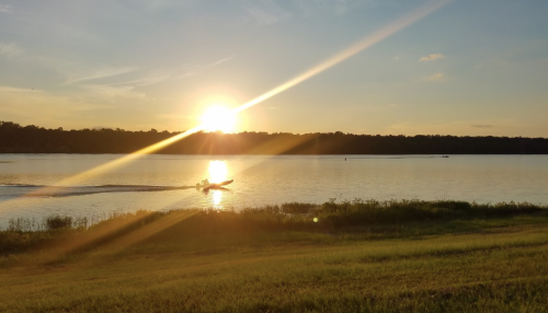 A serene sunset over a lake, with a boat gliding across the water and golden rays illuminating the scene.