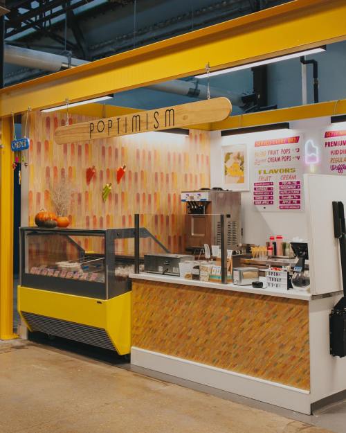 A colorful ice pop shop with a bright yellow counter, menu board, and a display case filled with treats.