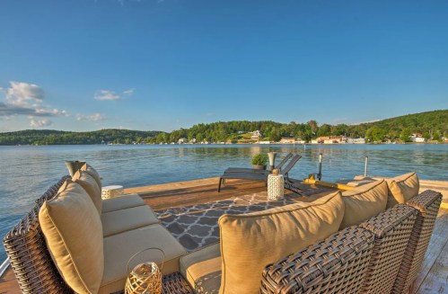 A cozy outdoor seating area overlooking a calm lake with green hills and houses in the background under a clear blue sky.