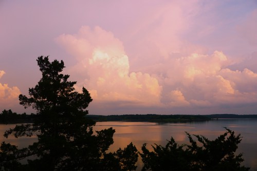 A serene lake at sunset, reflecting colorful clouds and surrounded by trees.