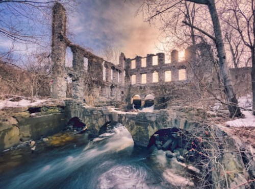 Ruins of a stone building beside a flowing stream, surrounded by trees and snow, under a colorful sky at sunset.