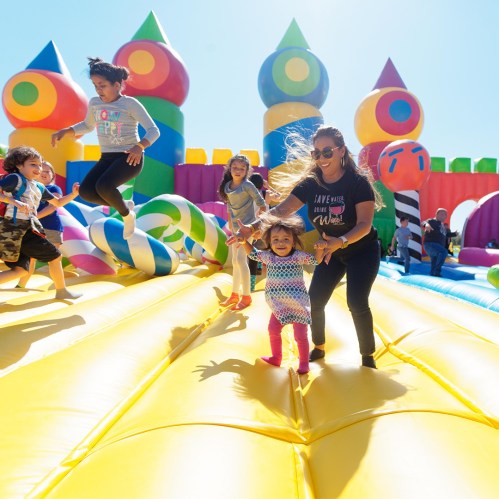 A joyful scene of children playing on a colorful bounce house with a smiling adult guiding them. Bright, sunny day.