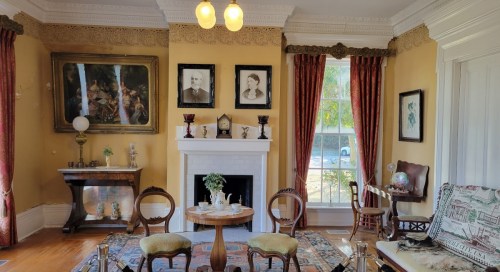 A vintage living room with a fireplace, antique furniture, framed portraits, and large windows letting in natural light.