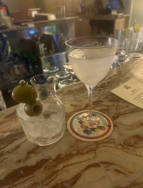 A cocktail glass with a clear drink and an olive, alongside a small glass of ice on a marble bar top.