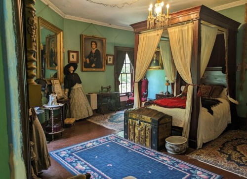 A vintage bedroom with a four-poster bed, ornate decor, and a figure in period clothing standing by a window.