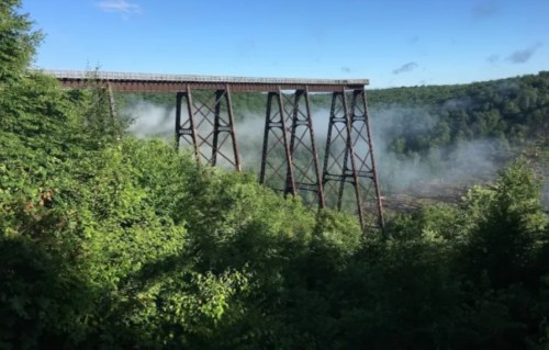 A tall, rusted bridge stands above a forested area, with mist rising from the ground and blue skies in the background.