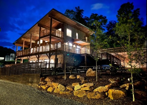 A modern wooden house at night, illuminated with warm lights, surrounded by trees and rocky landscaping.