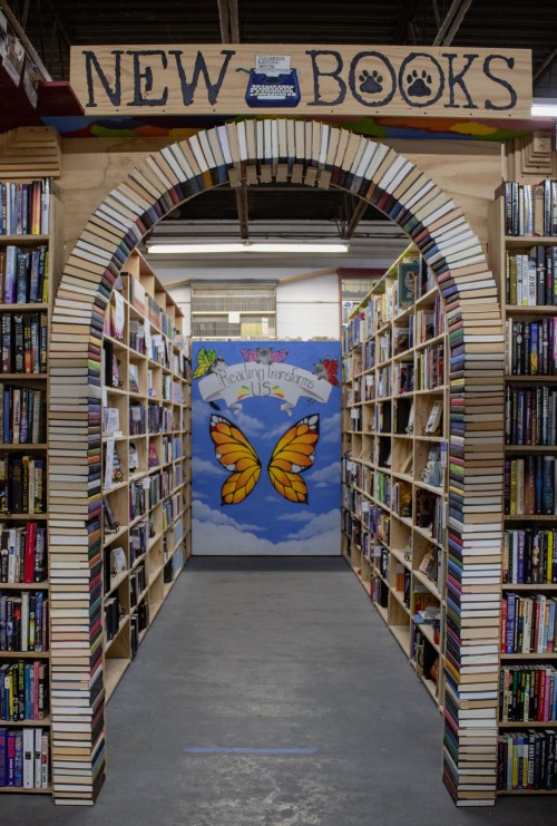 A colorful archway made of books leads into a cozy aisle of shelves filled with books, featuring a butterfly mural.