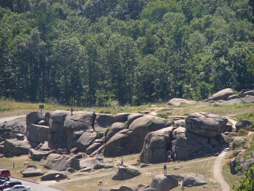A rocky outcrop with people exploring, surrounded by lush green trees and a winding path.