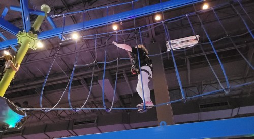 A person navigates a high ropes course, balancing on a narrow beam with safety gear in a brightly lit indoor space.
