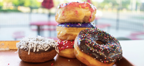 A stack of colorful donuts with sprinkles, frosting, and coconut on a tray, set against a blurred outdoor background.