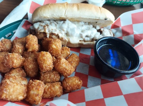 A sandwich with melted cheese and shredded meat, served with a side of crispy tater tots and a small cup of dipping sauce.