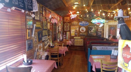 Cozy restaurant interior with wooden walls, vintage signs, and checkered tablecloths on tables. Warm, inviting atmosphere.