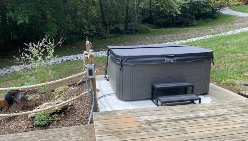 A hot tub with a cover sits on a concrete pad, surrounded by greenery and a wooden deck. Steps lead up to it.