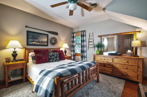 Cozy bedroom with a wooden bed, plaid bedding, lamps, and rustic decor, featuring a dresser and a window with natural light.