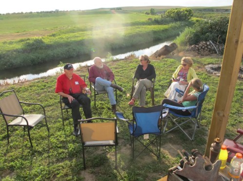 A group of four people sitting in chairs by a river, enjoying a conversation in a grassy outdoor setting.