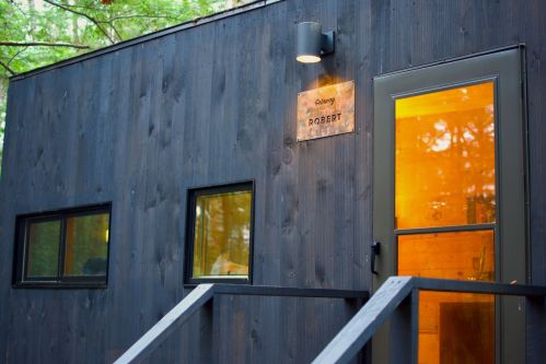 A modern cabin with dark wood siding, large windows, and a door labeled "Robert," surrounded by trees.