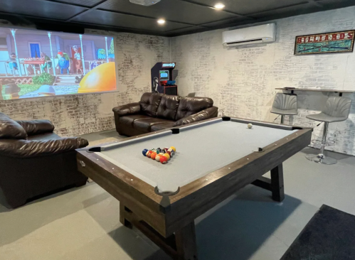 A cozy game room featuring a pool table, leather couches, a bar area, and a projector screen on the wall.