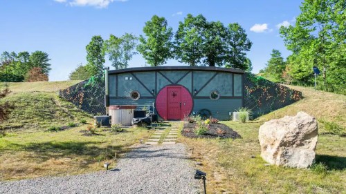 A unique, modern home with a red door, surrounded by greenery and a gravel pathway leading to it.