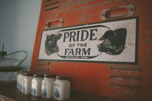 A vintage sign reading "Pride of the Farm" above small glass milk jars on a wooden shelf.