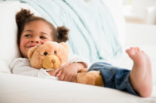 A smiling child cuddles a teddy bear while relaxing on a couch, with bare feet visible and a cozy atmosphere.