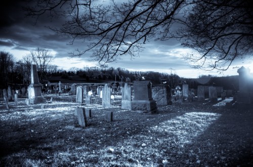A moody cemetery scene with gravestones under a cloudy sky, illuminated by soft light and shadows.