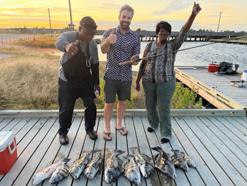 Three people stand on a dock, smiling and posing with fishing rods and a catch of fish at sunset.