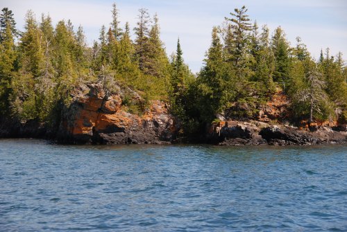 Rocky shoreline with lush green trees and calm blue water under a clear sky.