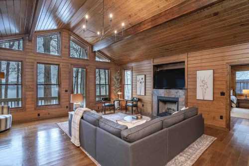 Cozy living room with wooden walls, large windows, a fireplace, and modern furniture overlooking a serene outdoor view.