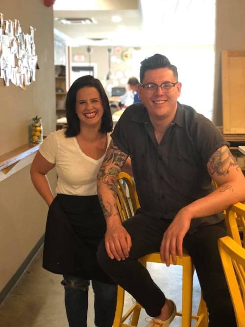 Two smiling individuals pose in a bright café, one standing and the other sitting on a yellow stool.